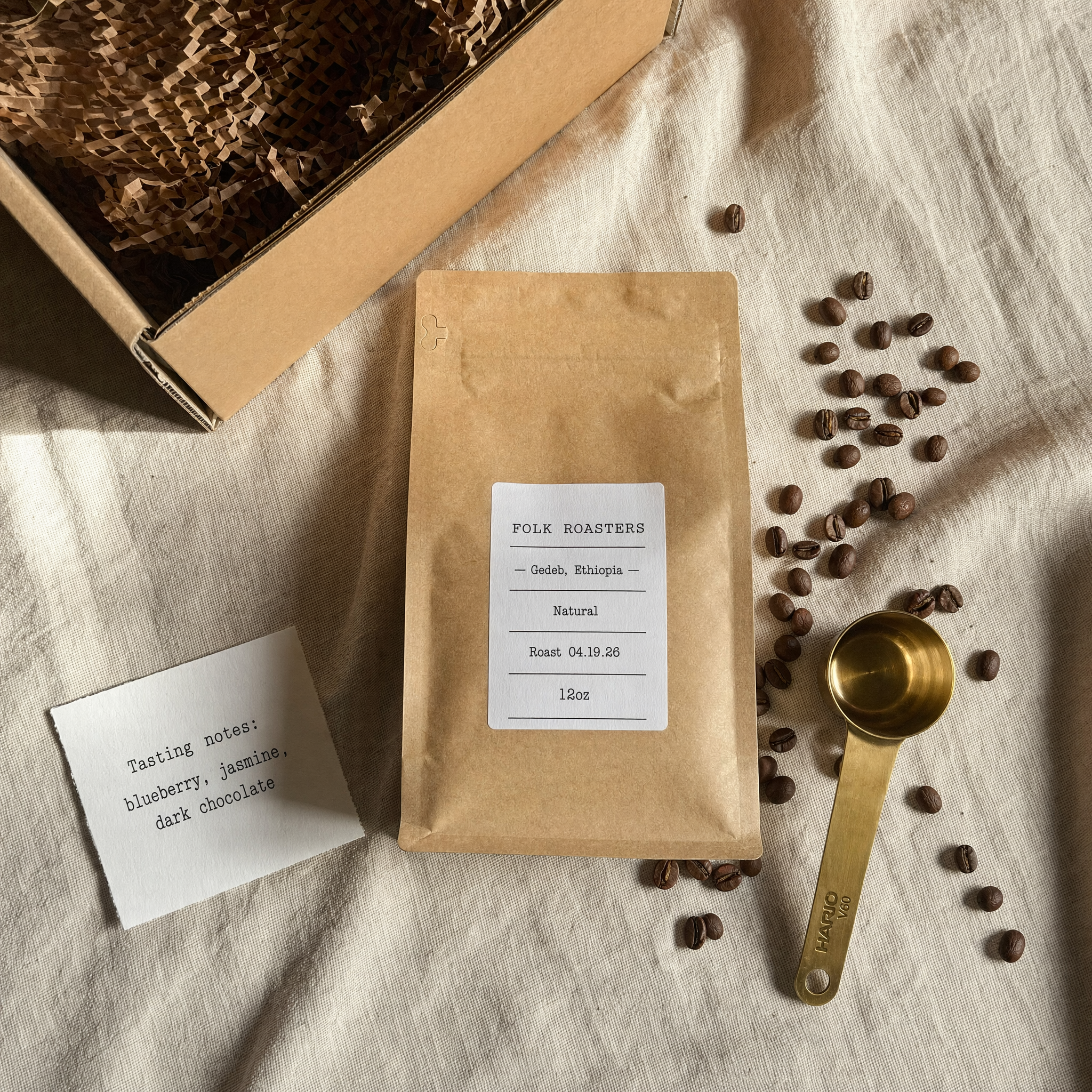 Overhead flatlay 4K, 4:5. Single-origin coffee subscription unboxing on unbleached linen: 250g kraft bag with exact label "FOLK ROASTERS — Gedeb, Ethiopia — Natural — Roast 04.19.26 — 12oz", brass Hario V60 scoop, small card reading exactly "Tasting notes: blueberry, jasmine, dark chocolate". Morning side-light, real coffee bean scatter, no color grade, no stock-photo steam.