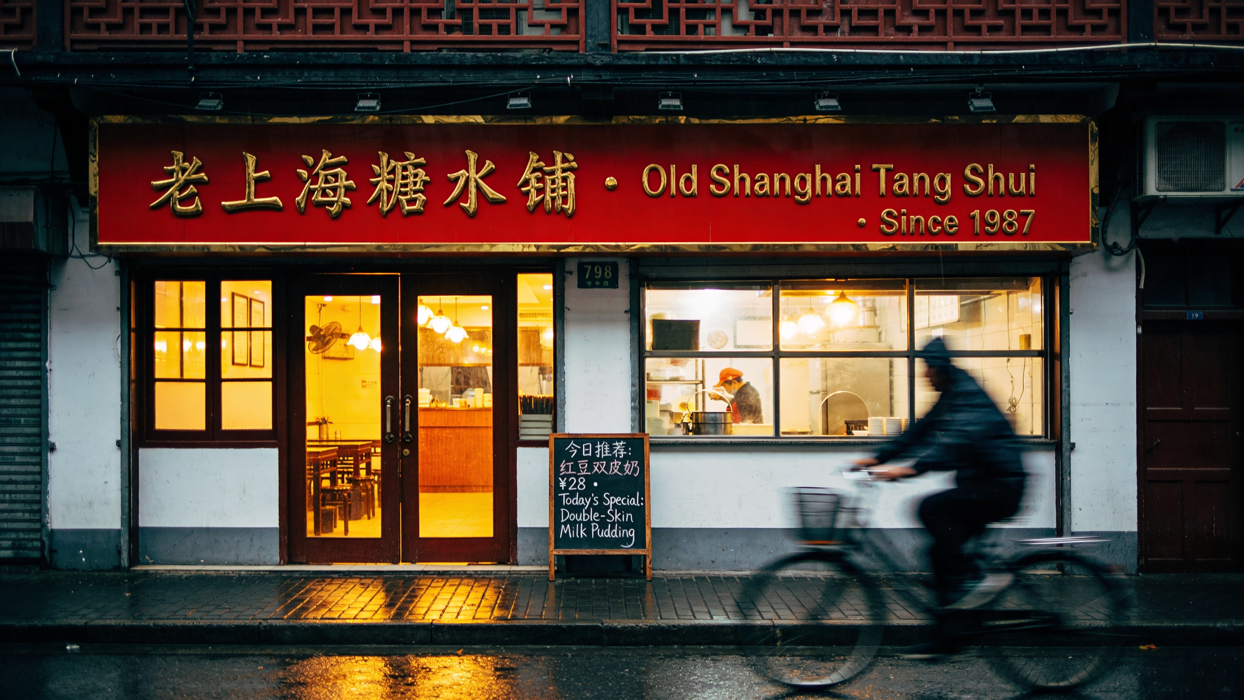 Shanghai Jing'an Temple Road storefront at 7 PM, rainy evening. Hand-painted bilingual sign above the door reads exactly "老上海糖水铺 · Old Shanghai Tang Shui · Since 1987" in red enamel with gold outline; below it a smaller chalkboard reads exactly "今日推荐：红豆双皮奶 ¥28 · Today's Special: Double-Skin Milk Pudding". Warm tungsten interior light spilling onto wet pavement, a cyclist in motion blur. Documentary photograph, 35mm f/2, Fujifilm Pro 400H look. Chinese and English both rendered verbatim, no swapped characters.