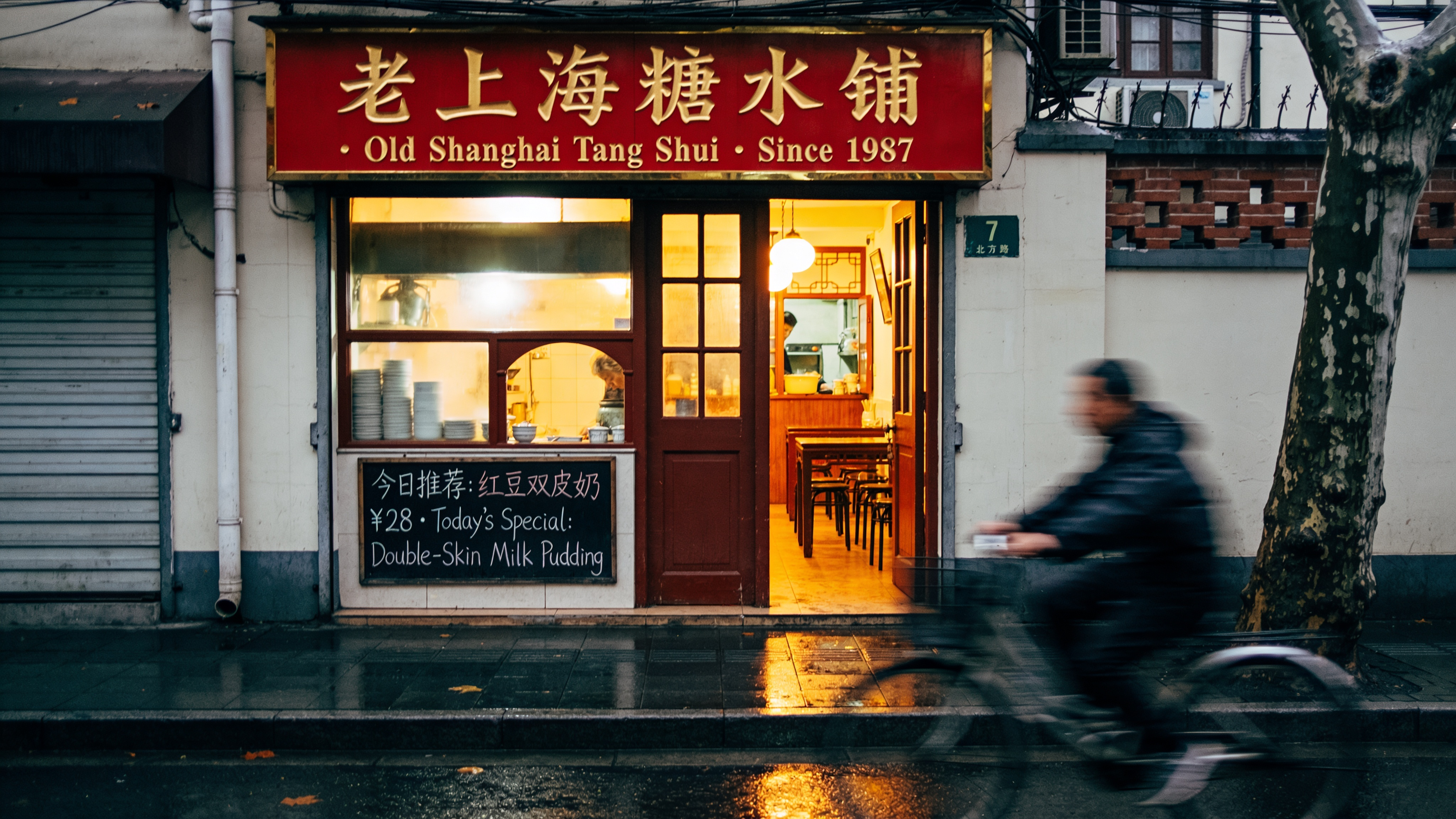 Shanghai Jing'an Temple Road storefront at 7 PM, rainy evening. Hand-painted bilingual sign above the door reads exactly "老上海糖水铺 · Old Shanghai Tang Shui · Since 1987" in red enamel with gold outline; below it a smaller chalkboard reads exactly "今日推荐：红豆双皮奶 ¥28 · Today's Special: Double-Skin Milk Pudding". Warm tungsten interior light spilling onto wet pavement, a cyclist in motion blur. Documentary photograph, 35mm f/2, Fujifilm Pro 400H look. Chinese and English both rendered verbatim, no swapped characters.