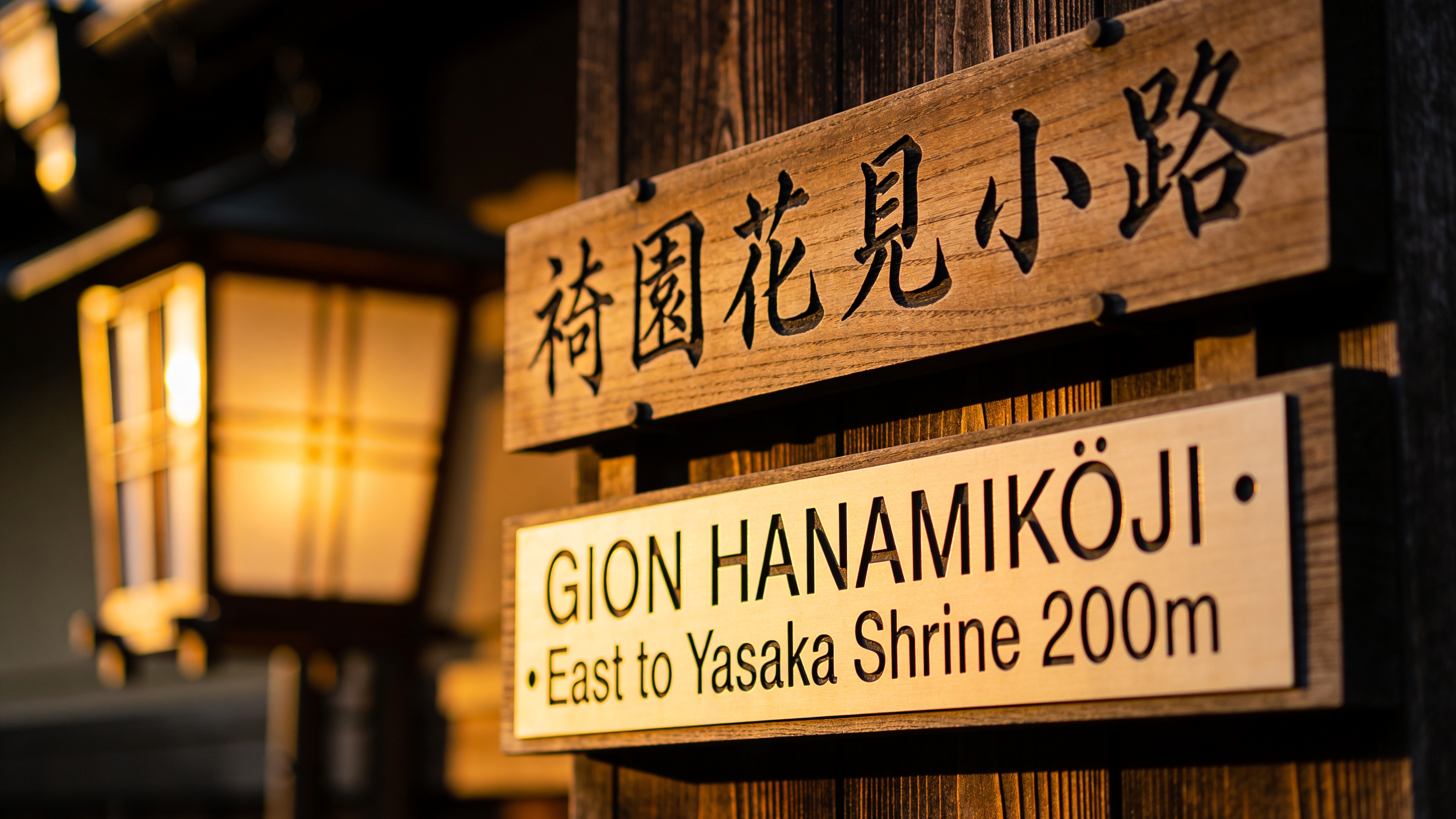 Kyoto Gion bilingual street sign mounted on dark wood, golden-hour backlight. Vertical sign top reads exactly "祇園花見小路" in brushstroke calligraphy; below horizontally in sans serif reads exactly "GION HANAMIKŌJI · East to Yasaka Shrine 200m". Subtle wood grain, a single lantern out of focus. Shot on Leica Q2 28mm f/2. Japanese characters and English rendered verbatim, correct kerning.