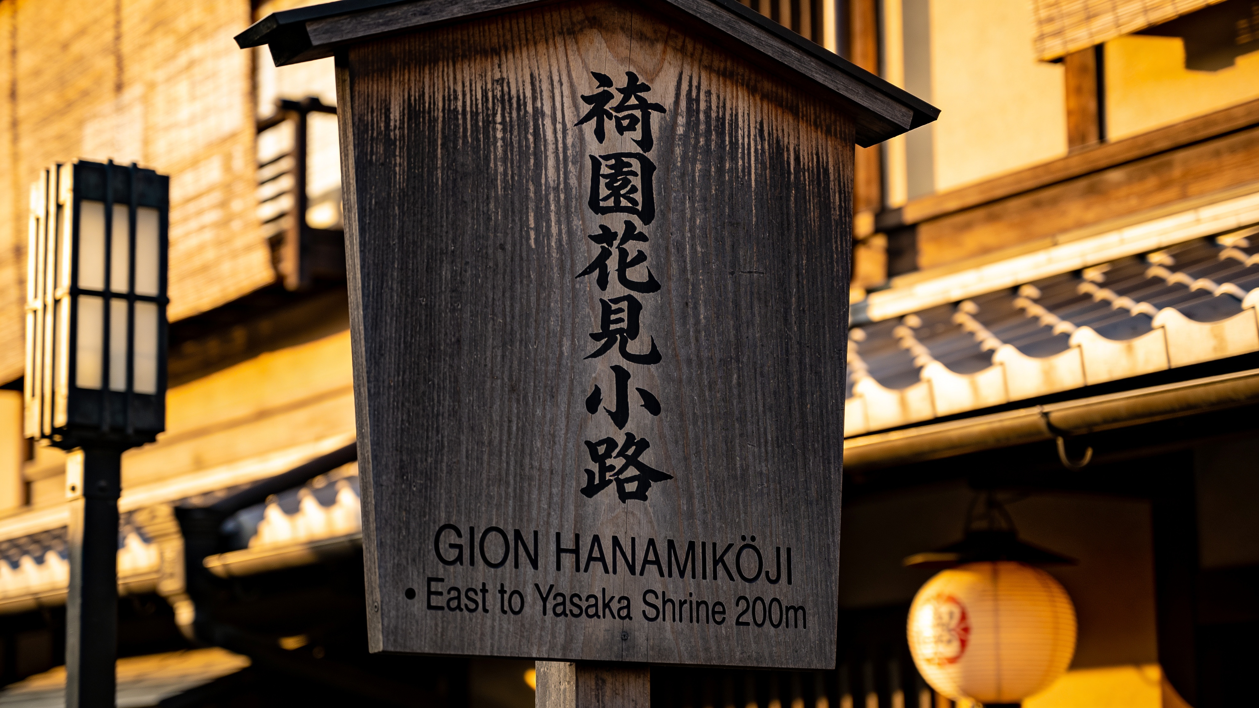 Kyoto Gion bilingual street sign mounted on dark wood, golden-hour backlight. Vertical sign top reads exactly "祇園花見小路" in brushstroke calligraphy; below horizontally in sans serif reads exactly "GION HANAMIKŌJI · East to Yasaka Shrine 200m". Subtle wood grain, a single lantern out of focus. Shot on Leica Q2 28mm f/2. Japanese characters and English rendered verbatim, correct kerning.