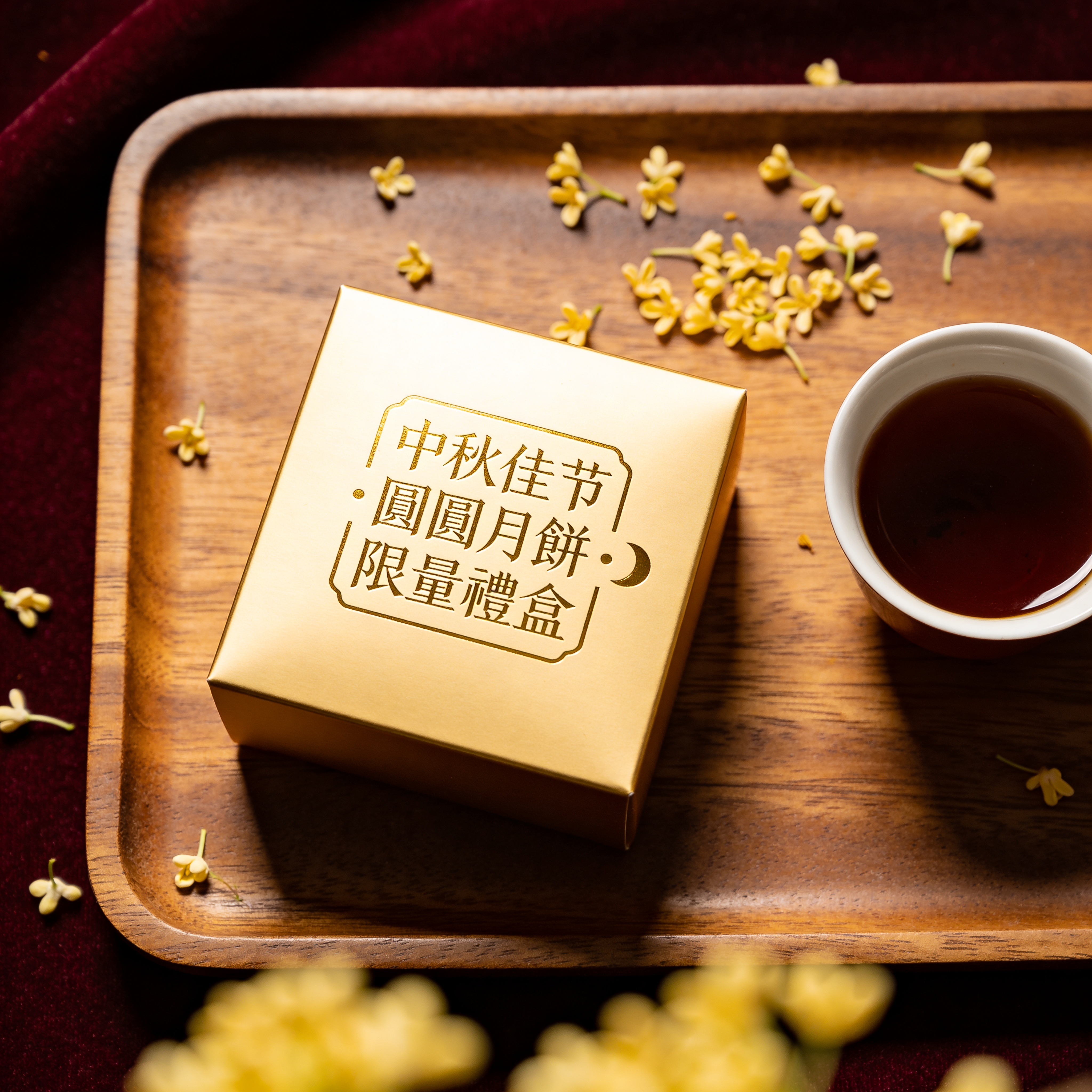 Moon cake box packshot, overhead flatlay on walnut tray. Square hot-stamped gold lid against deep burgundy fabric, lid text reads exactly "中秋佳节 · 團圓月餅 · 限量禮盒" in traditional seal-script style with a small crescent icon. Loose osmanthus blossoms scattered on tray, a single cup of dark oolong to the right. Editorial product photo, 80mm macro, f/8 focus-stacked. Simplified characters rendered exactly, gold foil texture visible on characters.