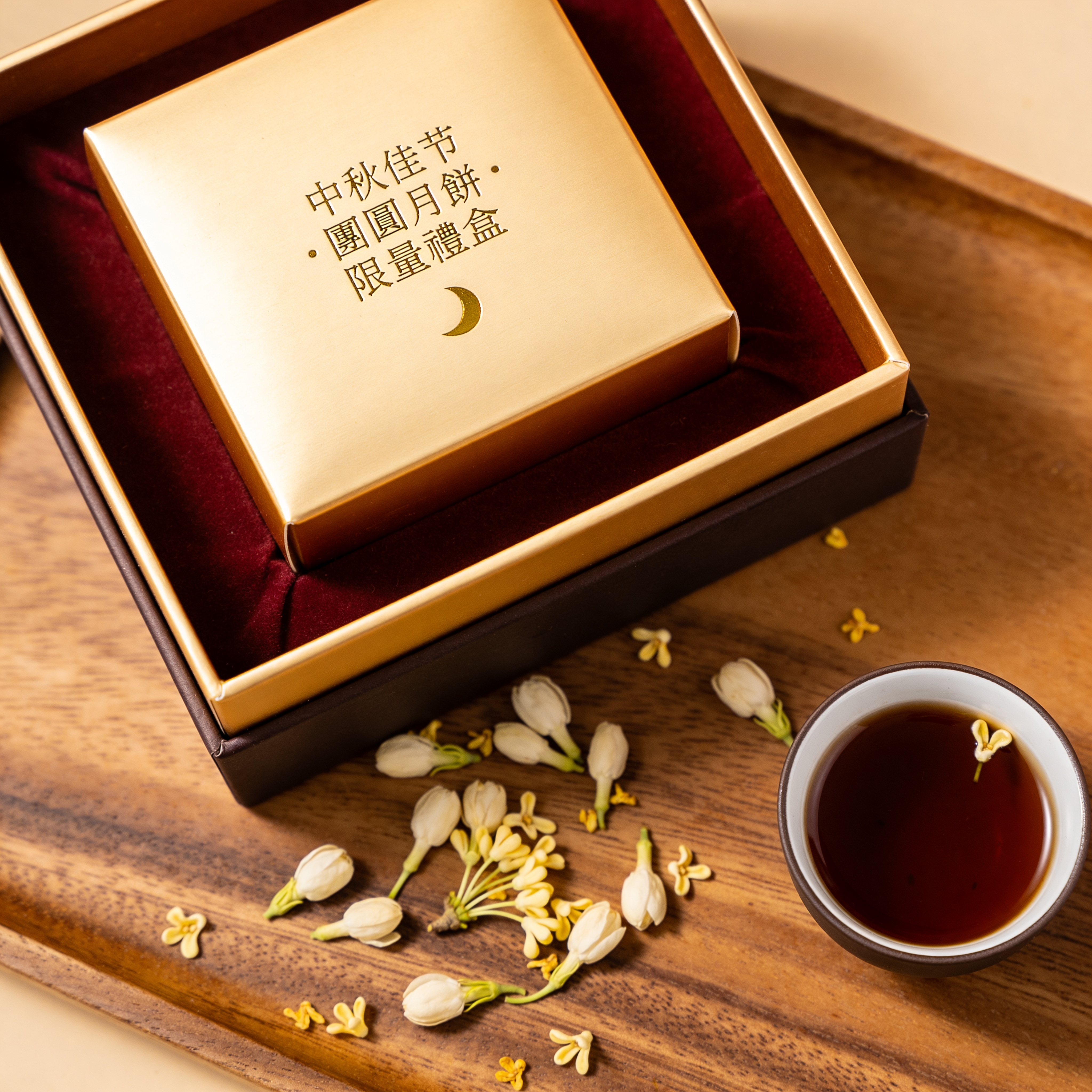 Moon cake box packshot, overhead flatlay on walnut tray. Square hot-stamped gold lid against deep burgundy fabric, lid text reads exactly "中秋佳节 · 團圓月餅 · 限量禮盒" in traditional seal-script style with a small crescent icon. Loose osmanthus blossoms scattered on tray, a single cup of dark oolong to the right. Editorial product photo, 80mm macro, f/8 focus-stacked. Simplified characters rendered exactly, gold foil texture visible on characters.
