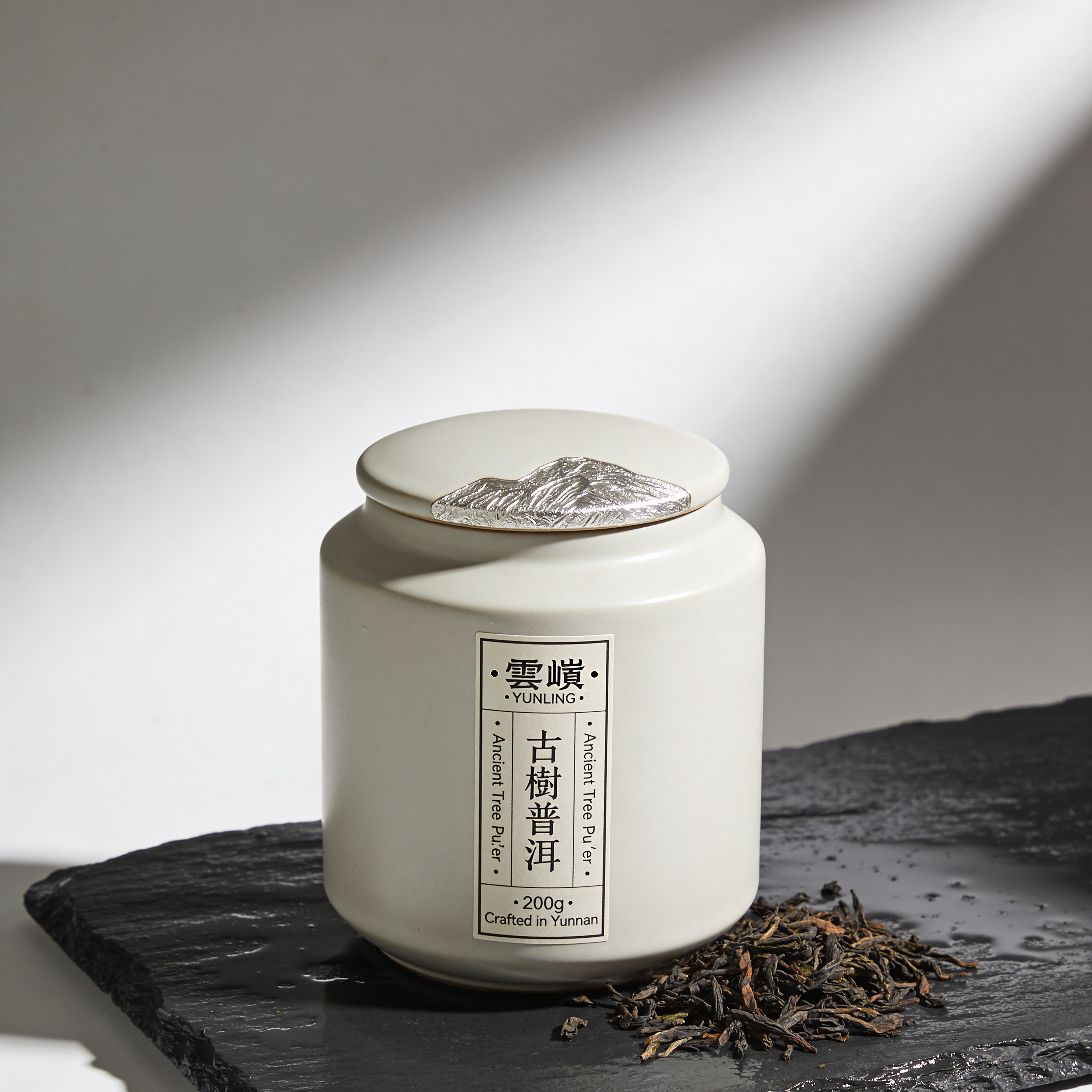Hero packshot for a fictional Chinese premium tea brand. Matte-ceramic canister standing centered on rain-dark slate. Front label in vertical bilingual layout reads exactly "雲嶺 · YUNLING · Ancient Tree Pu'er · 古樹普洱 · 200g · Crafted in Yunnan". Silver-leaf accents on the lid, loose tea leaves scattered at base. Editorial side light at 45°, Hasselblad X2D 80mm f/8, focus-stacked sharp, pure soft shadow. Chinese characters and English rendered verbatim, no typo.