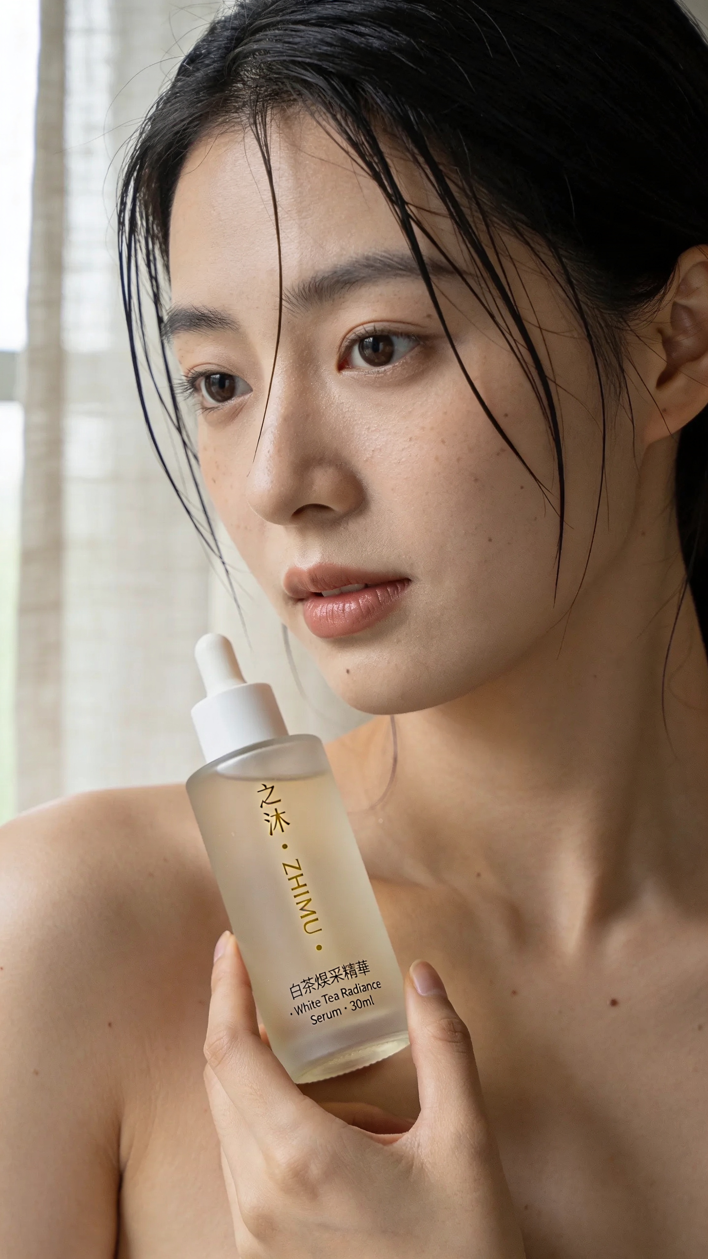 Editorial lifestyle shot for a fictional Shanghainese skincare brand. A young Asian woman, 28, with bare shoulders and damp hair, holds a frosted glass serum bottle near her collarbone. Bottle label reads exactly "之沐 · ZHIMU · 白茶煥采精華 · White Tea Radiance Serum · 30ml" in vertical gold leaf. Soft north-window light, linen curtain out of focus. Portra 400 palette, skin pores visible, no beauty retouch. Chinese and English both rendered correctly.