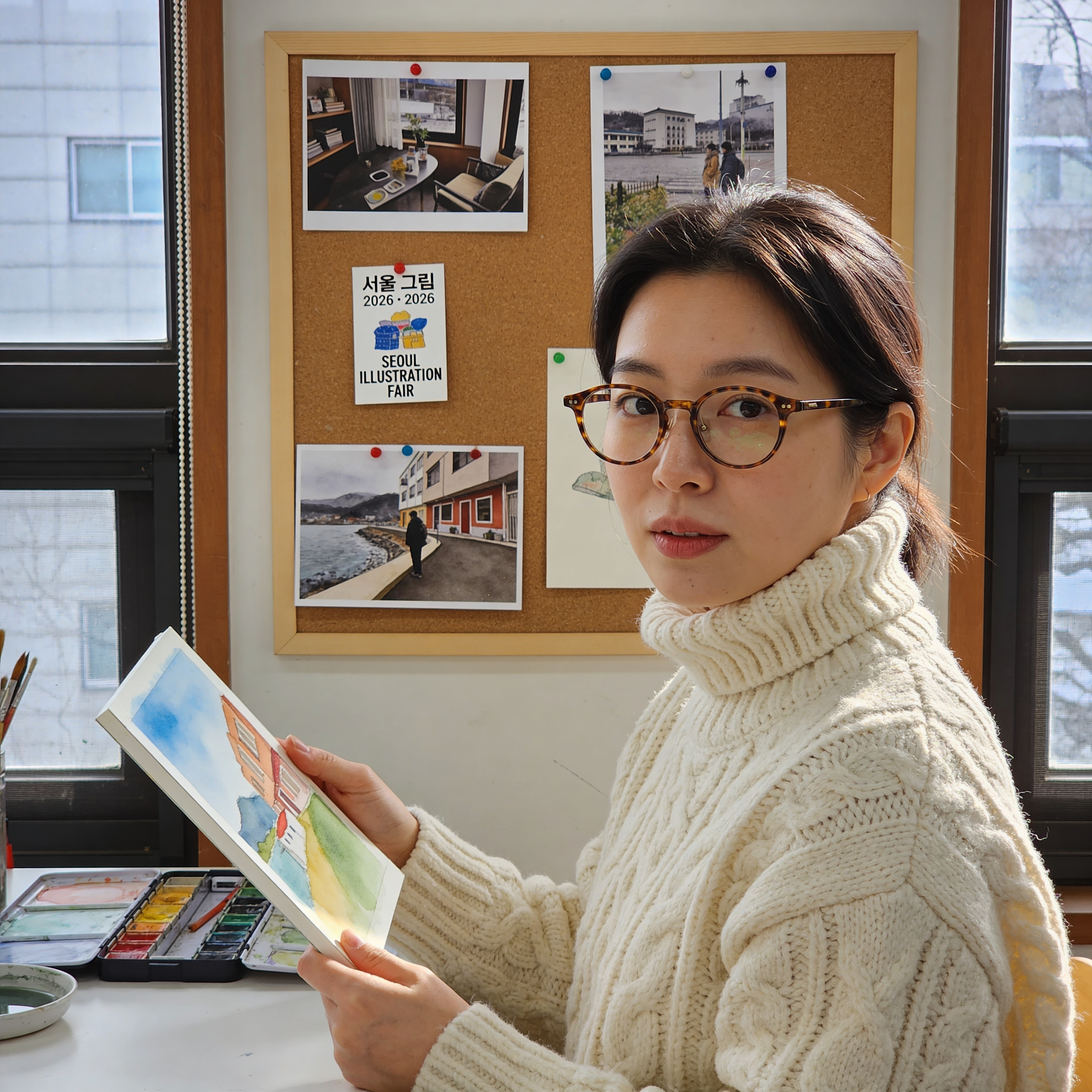 Half-length portrait of a 31-year-old Korean illustrator at her Seoul studio, winter afternoon. She's wearing a chunky cream-colored knit and round tortoise-shell glasses, holding a half-finished watercolor. Bulletin board behind her pinned with inspiration clippings; one small postcard on it reads exactly "서울 그림 2026 · SEOUL ILLUSTRATION FAIR". Sony A7IV 55mm f/1.8, window light, honest skin detail, no filter look.